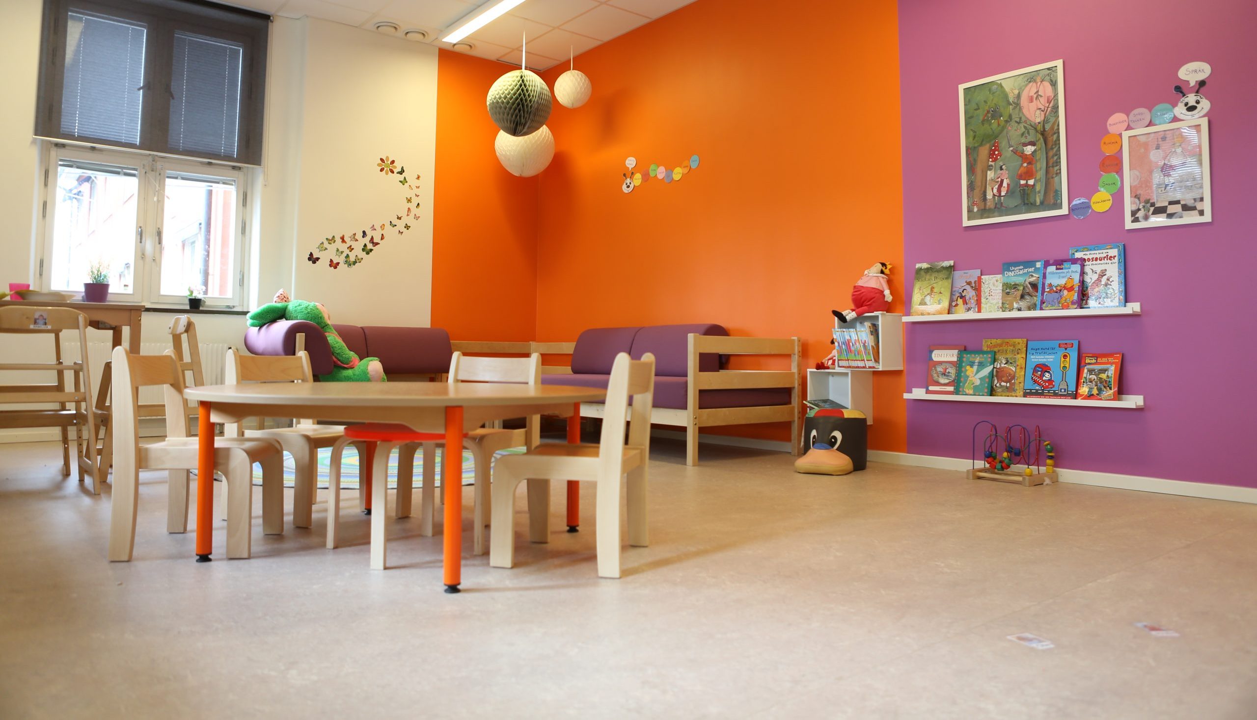 Färgglatt rum på förskola med orange och rosa väggar, en läshörna med bokhyllor fulla av barnböcker, och en sittgrupp med lila soffor och ett runt bord med träpallar. Vita pappersbollar hänger i taket och fjärilsklistermärken pryder väggen.