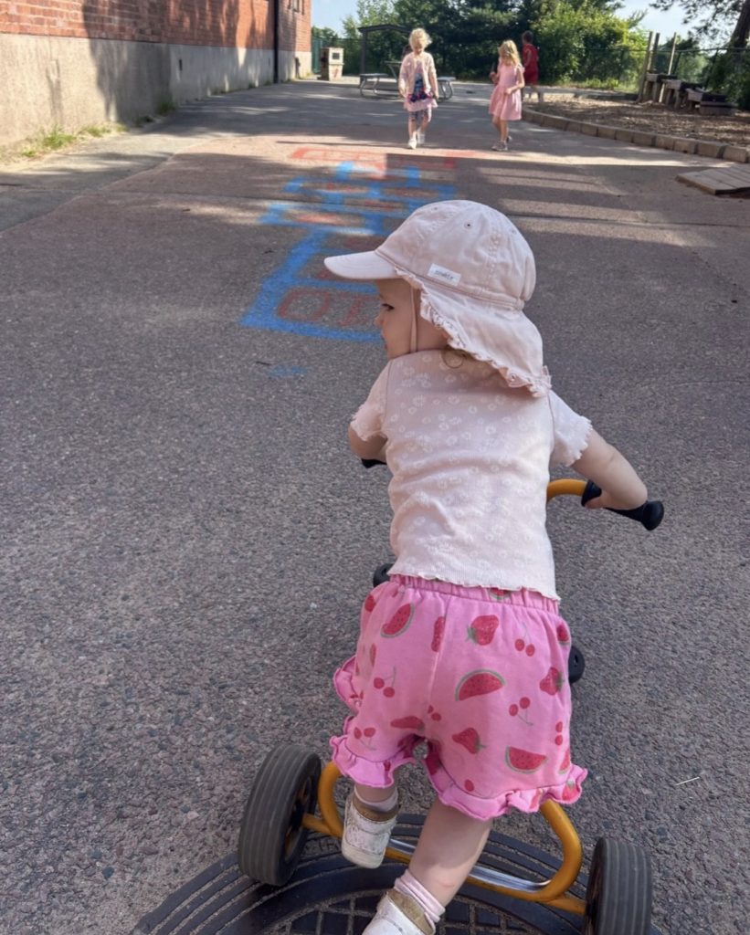 Ett litet barn i beige solhatt och rosa shorts med frukt-mönster kör en gul trehjuling på en asfalterad skolgård, med hopprutor målade på marken och andra barn som leker i bakgrunden.