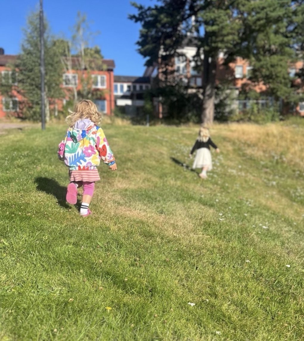 Två barn springer på en grönskande gräsmatta en solig dag. Barnet i förgrunden bär en färgglad blommig jacka, medan det andra barnet syns längre bort i bakgrunden. Tegelbyggnader och träd syns i horisonten under en klarblå himmel.