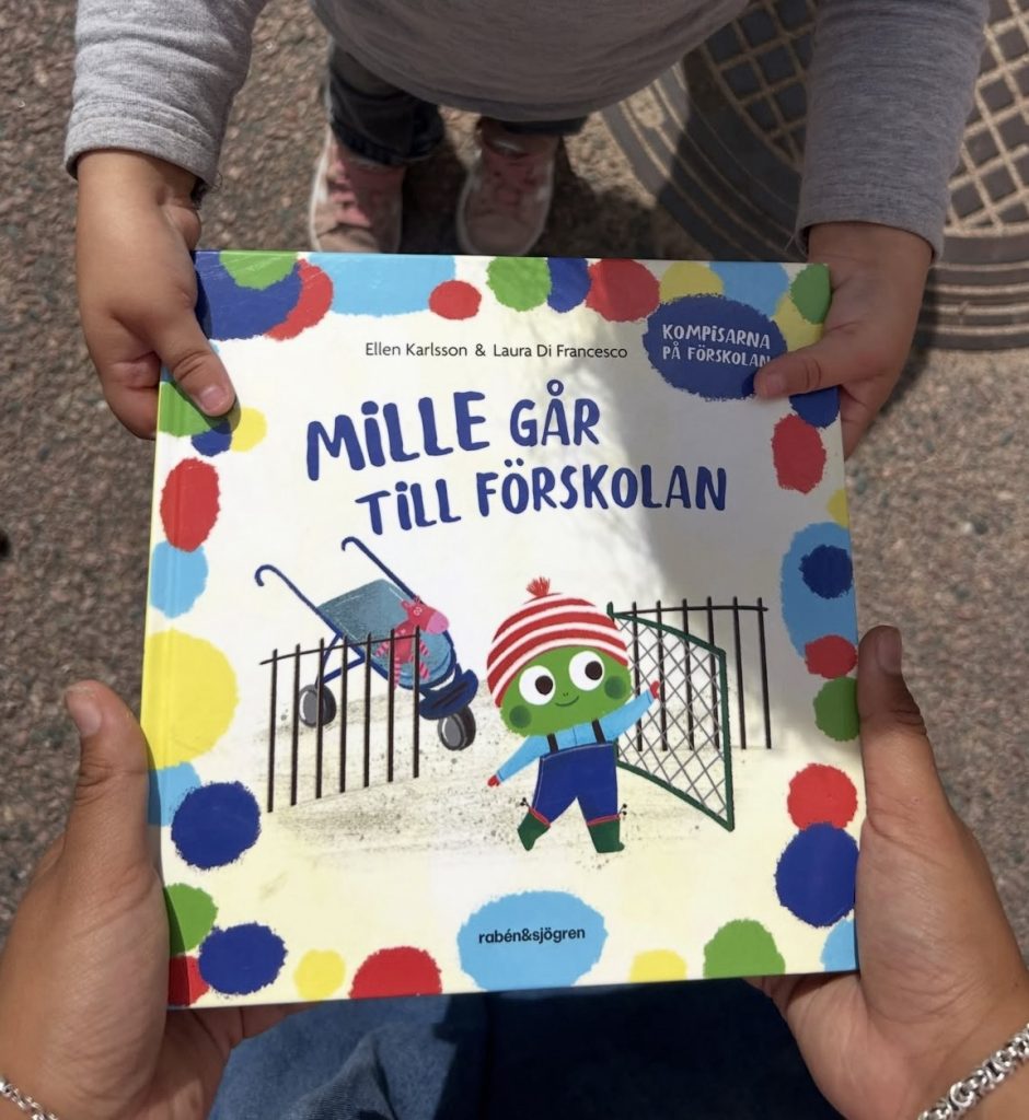 Ett barn håller upp barnboken 'Mille går till förskolan' av Ellen Karlsson och Laura Di Francesco, med ett färgglatt omslag som visar en tecknad grön figur i mössa vid en förskolegård.