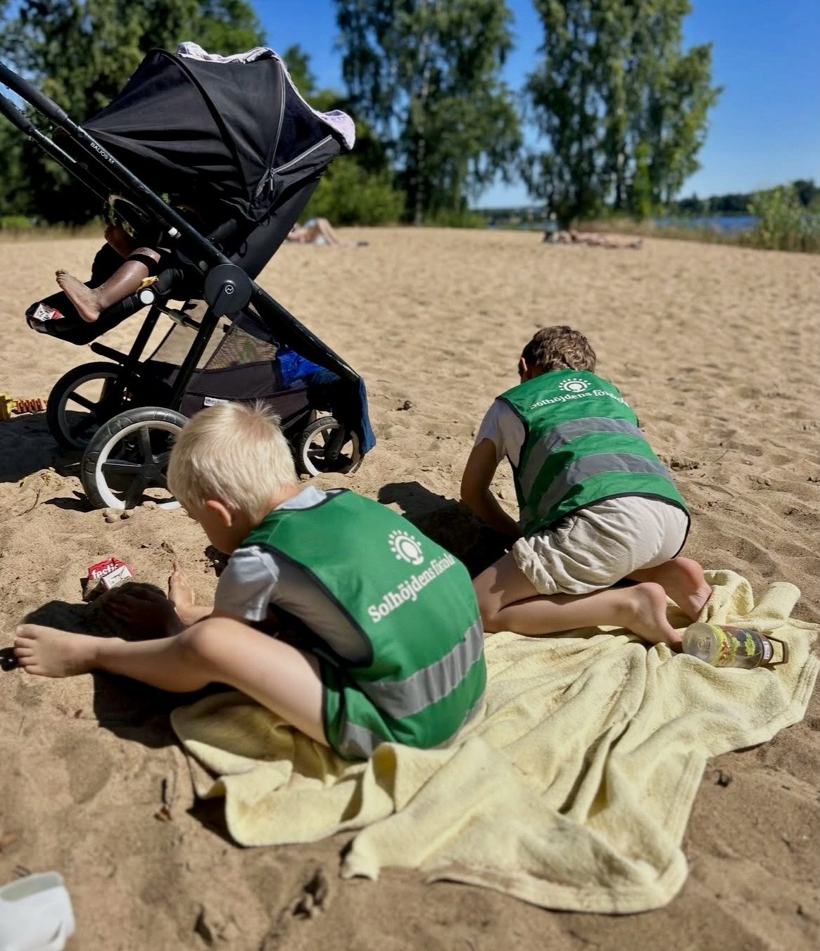 Två barn i gröna reflexvästar från 'Solhöjdens förskola' leker och gräver i sanden på en solig badstrand, liggandes på en gul handduk. En barnvagn står bredvid, och träd samt vatten syns i bakgrunden.