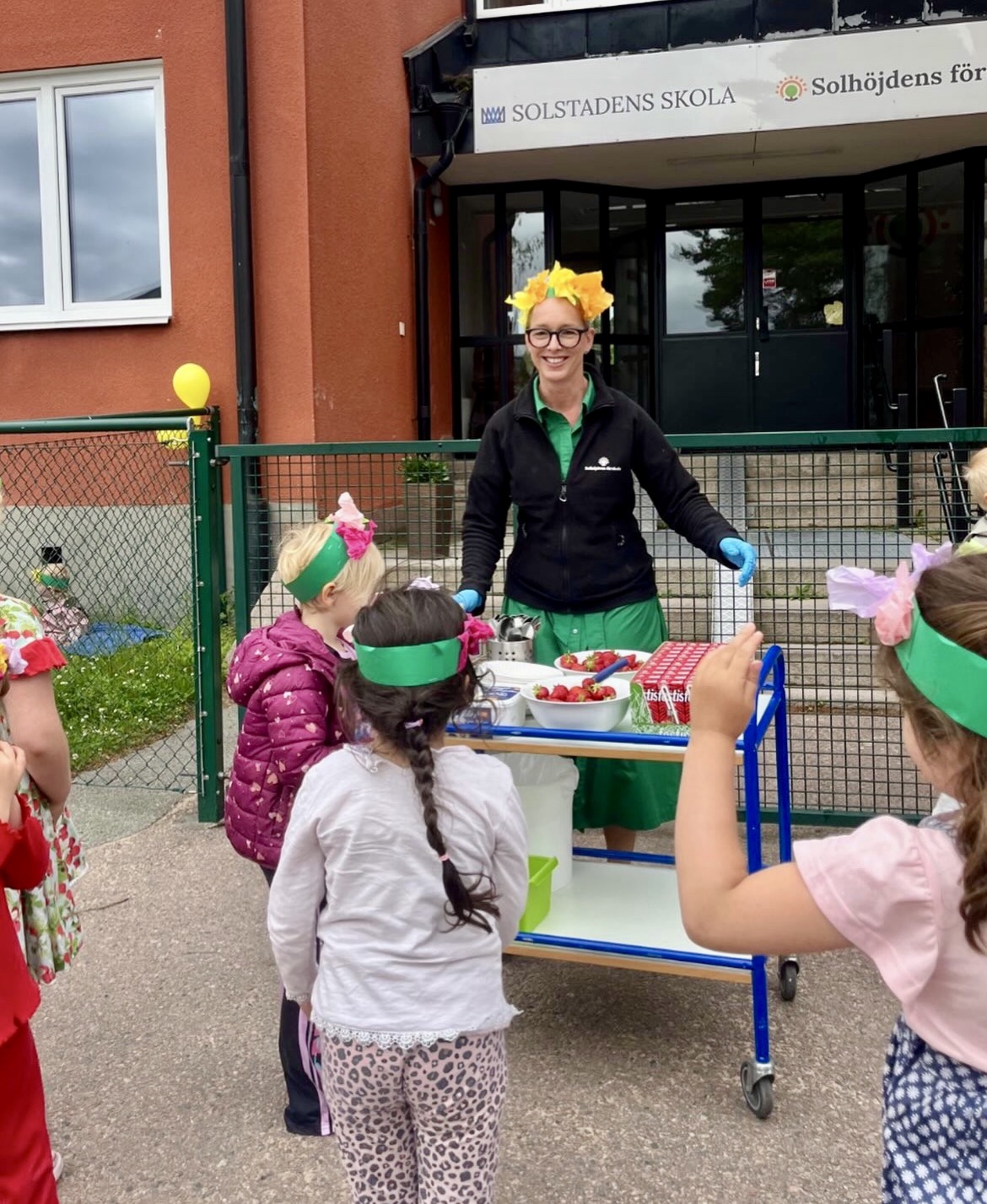 En leende vuxen med blomsterkrans på huvudet serverar jordgubbar från en vagn till en grupp barn utomhus vid skylten 'Solstadens skola / Solhöjdens förskola'. Flera barn bär gröna pannband med blommor, troligtvis inför en midsommarfirande.