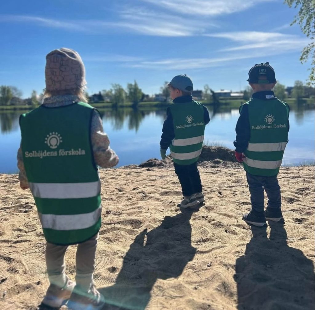 Tre barn i gröna reflexvästar med texten 'Solhöjdens förskola' står på en sandstrand och blickar ut över en lugn sjö under en klarblå himmel med lätta moln
